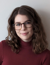 Reba looks directly into the camera, smiling. She has shoulder length, wavy brown hair. She has brown tortoise-shell glasses, and is wearing a maroon turtle neck.