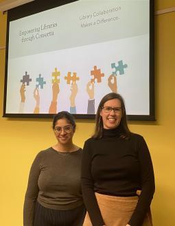 Sameena and Leah stand in front of a screen with the first slide of their presentation showing. Both are smiling at the camera.