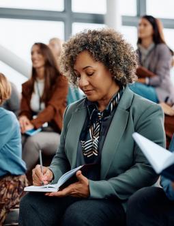 A woman takes notes in a small notebook at the front of an audience. 